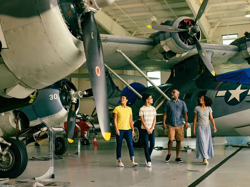 A family of four walk together inside an aircraft hangar at the Military Aviation Museum, with large vintage military planes in the background.