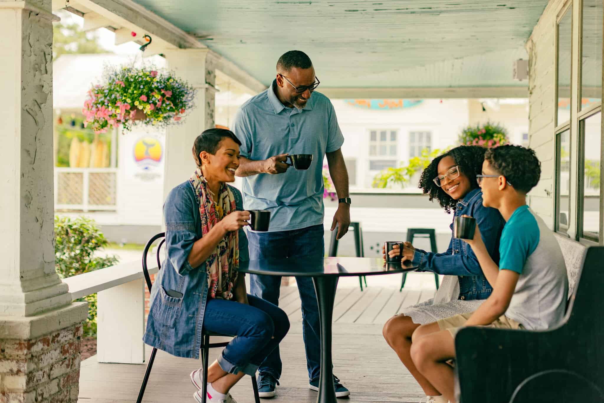 Four people sit and stand around a table on a porch, smiling and holding coffee mugs, with hanging plants and a house in the background.