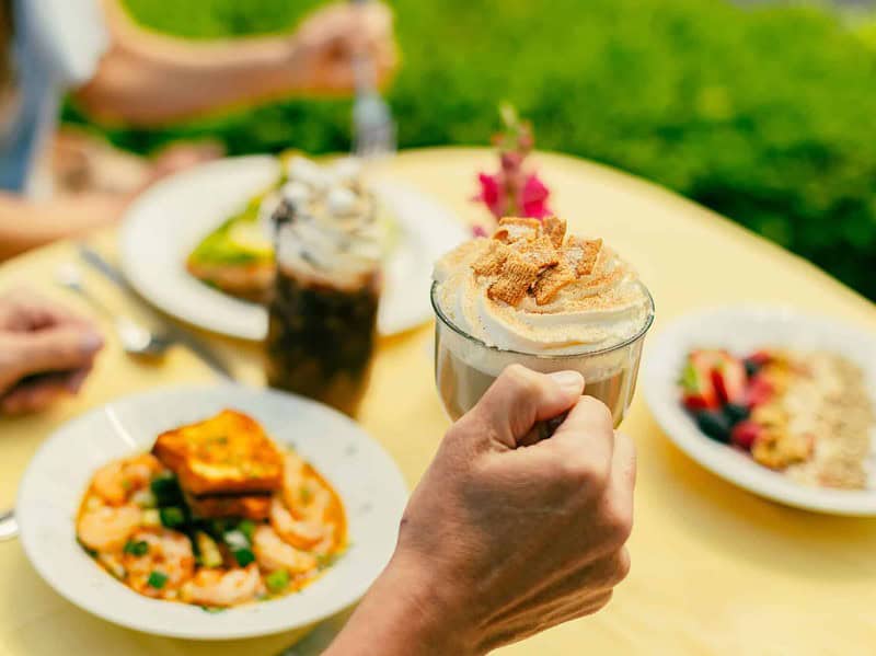 A hand holds a glass of coffee topped with whipped cream. Plates of breakfast food and another drink are on a yellow outdoor table at The Bee and The Biscuit restaurant.