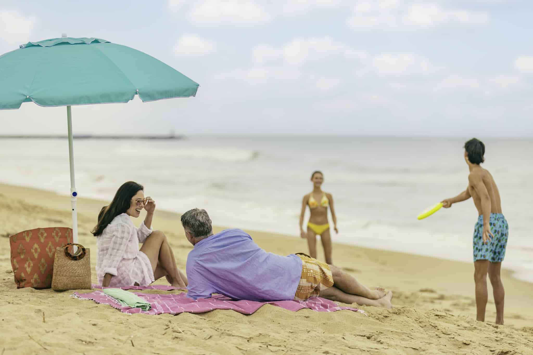 Two adults relax on a blanket under an umbrella on Croatan Beach while two young people play with a frisbee near the shore.