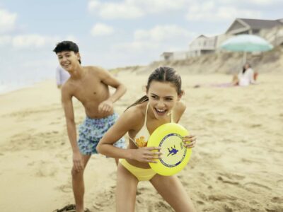 A girl in a yellow swimsuit holds a yellow frisbee while smiling on a sandy beach, with a boy in swim trunks standing behind her. Houses and a beach umbrella are in the background.