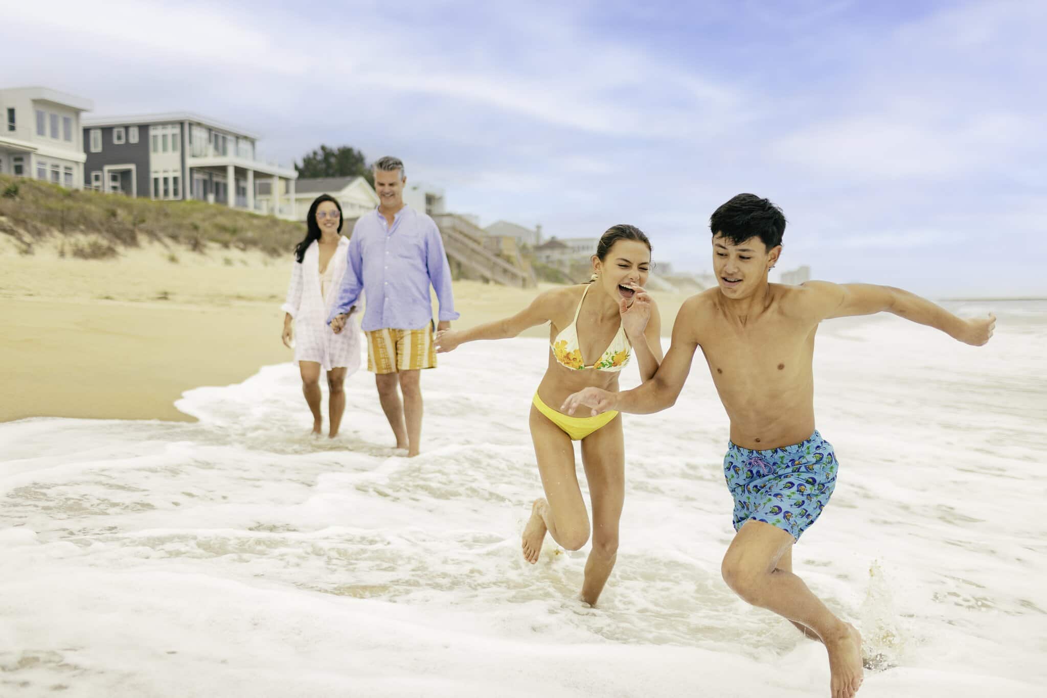 Two adults walk along the beach while a young man and woman in swimsuits play in the waves near the shoreline on a cloudy day.