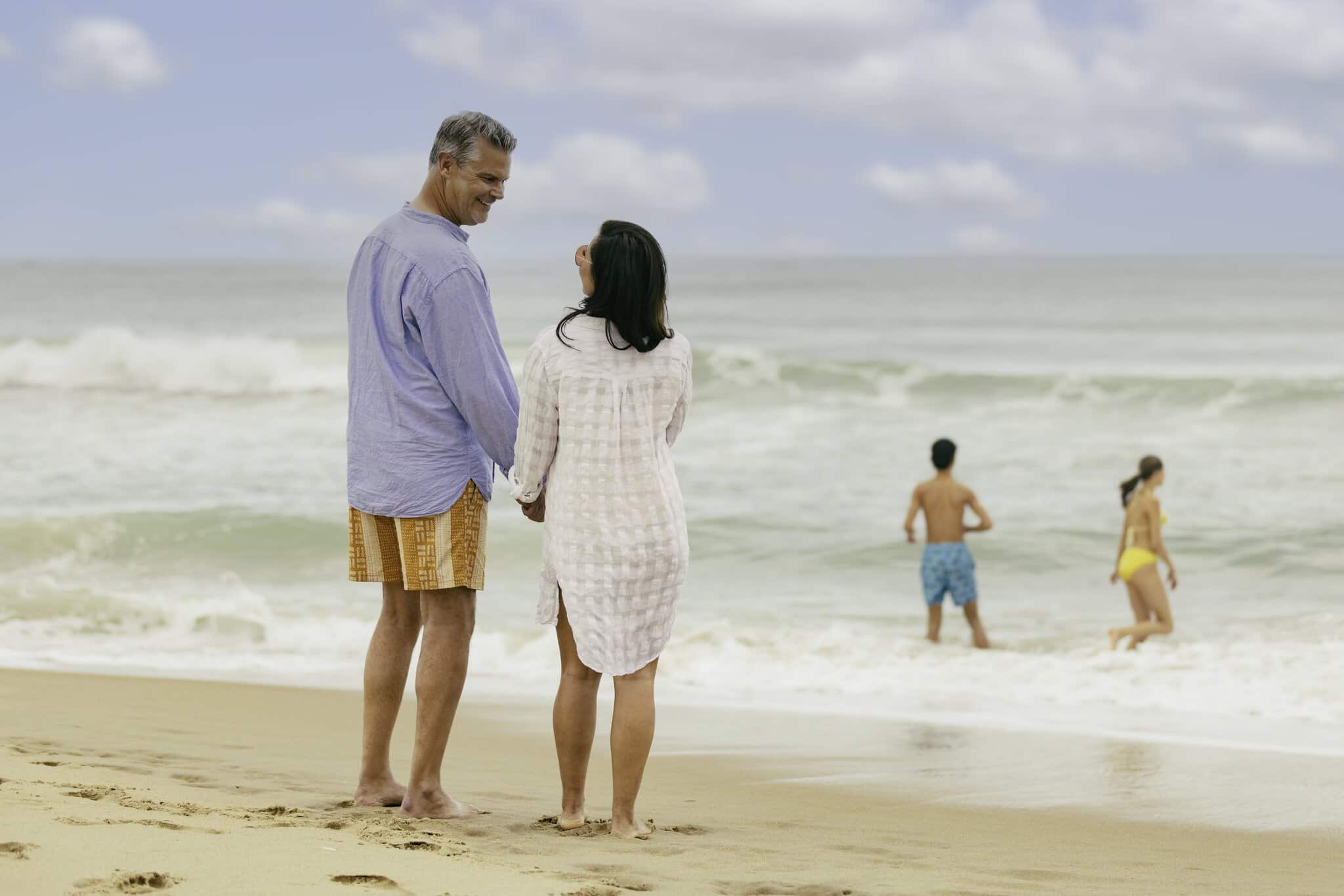 A man and woman stand holding hands on a sandy beach, facing the ocean, while two people swim in the water nearby.