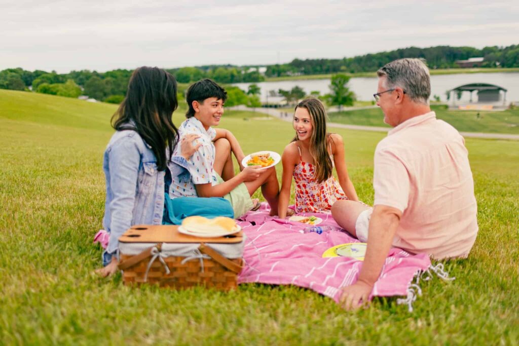 Four people sit on a pink blanket having a picnic with food and a basket on a grassy hill at Mount Trashmore near a lake, smiling and talking on a cloudy day.