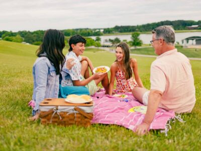 Four people sit on a pink blanket having a picnic with food and a basket on a grassy hill near a lake, smiling and talking on a cloudy day.