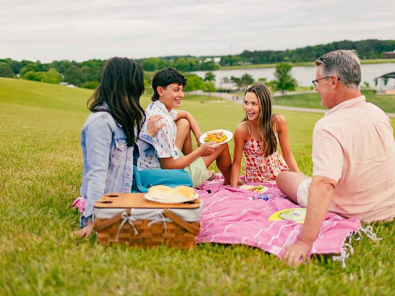 Four people sit on a pink blanket having a picnic with food and a basket on a grassy hill at Mount Trashmore near a lake, smiling and talking on a cloudy day.