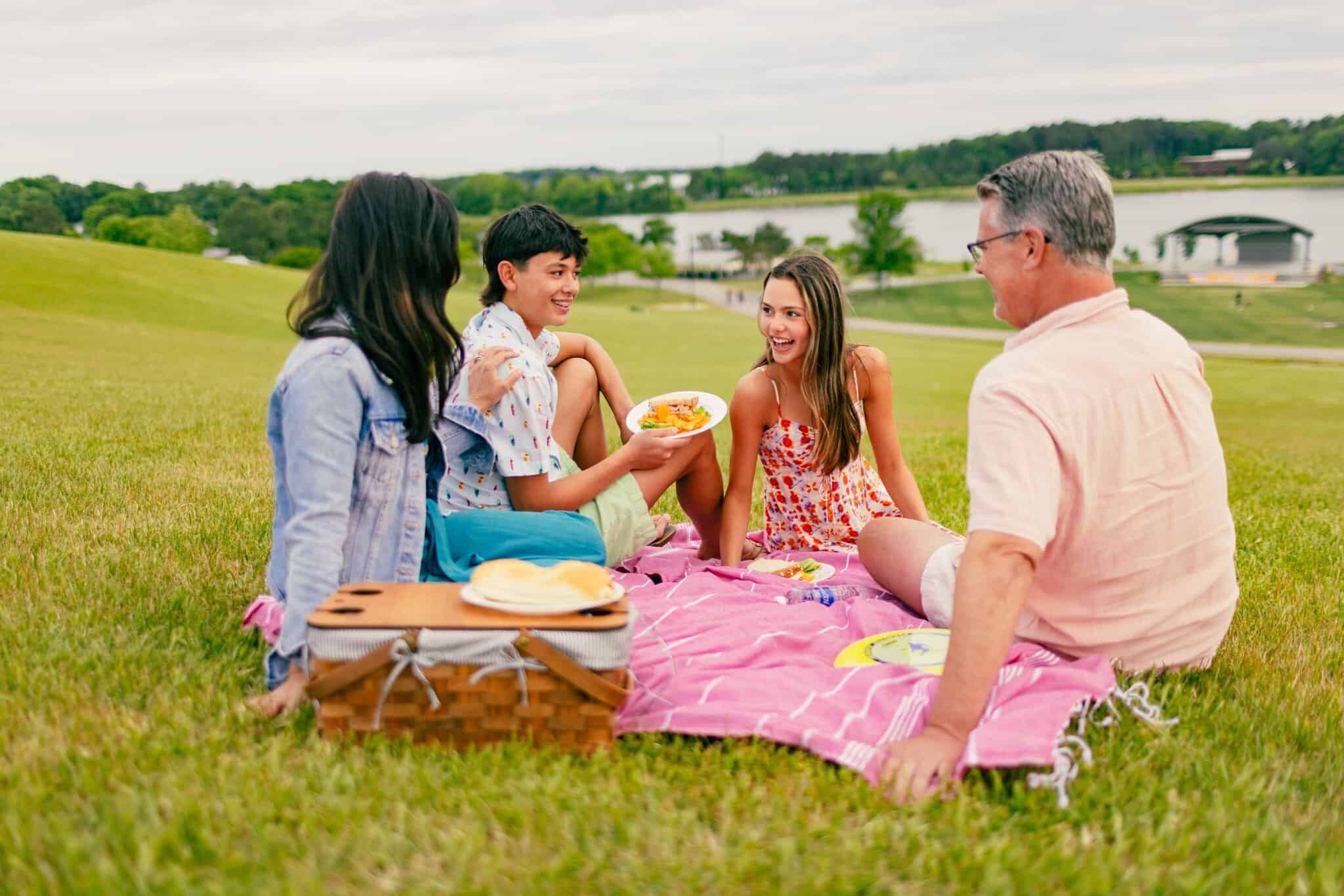 Four people sit on a pink blanket having a picnic with food and a basket on a grassy hill near a lake, smiling and talking on a cloudy day.