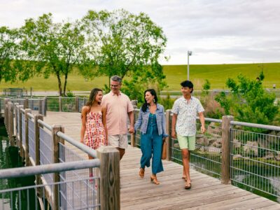 Four people walk together on a wooden boardwalk over water, surrounded by trees and grassy hills, on a partly cloudy day.