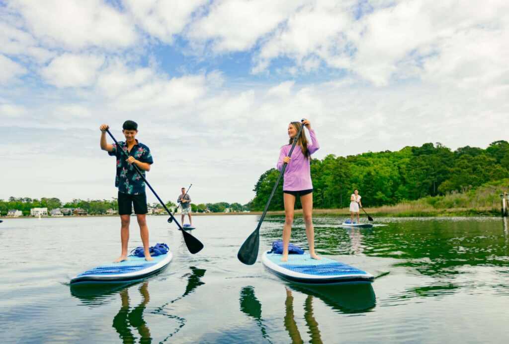 Five people paddleboarding on calm water, surrounded by greenery under a partly cloudy sky.