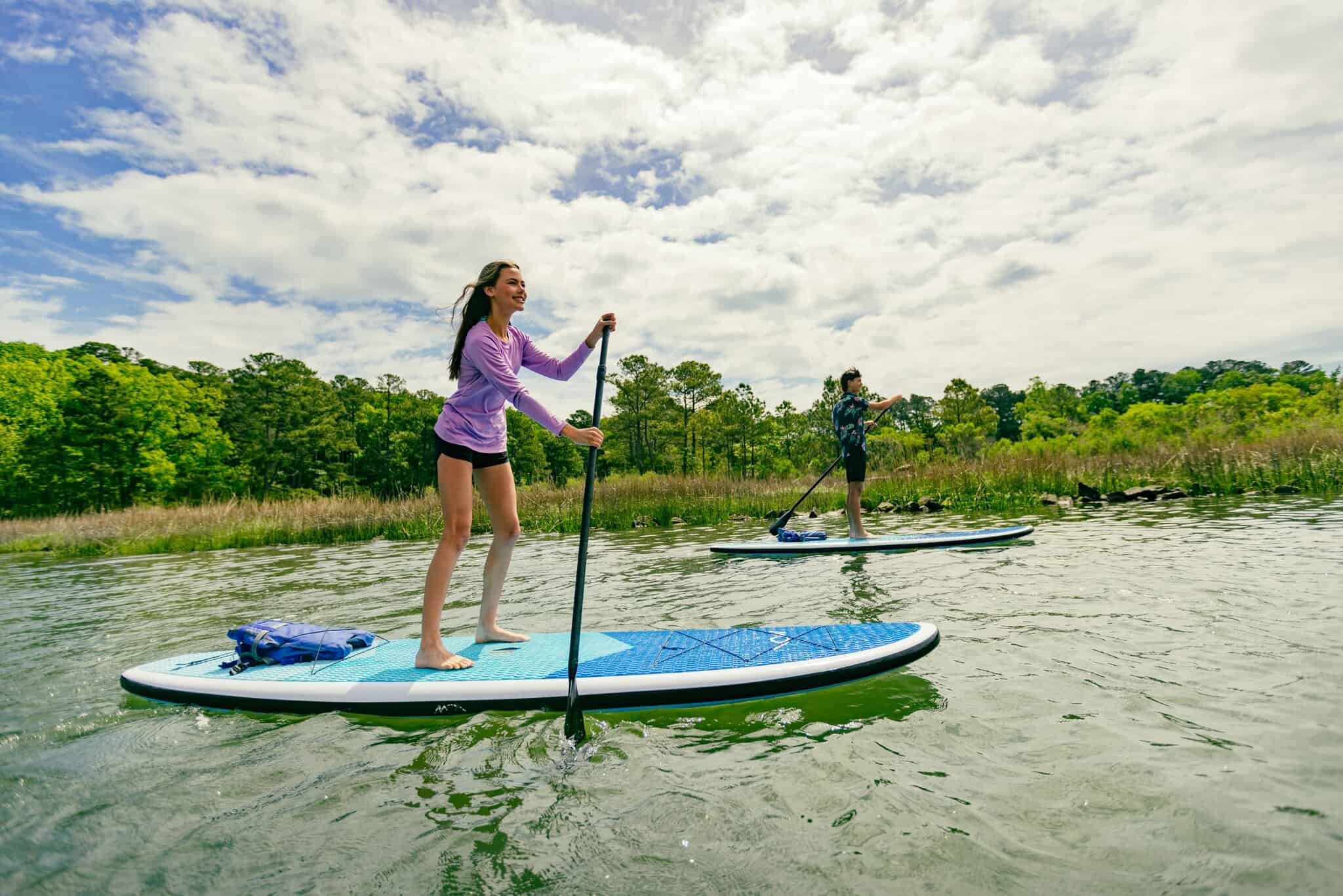 Two people paddleboarding on a calm body of water, surrounded by greenery under a partly cloudy sky.