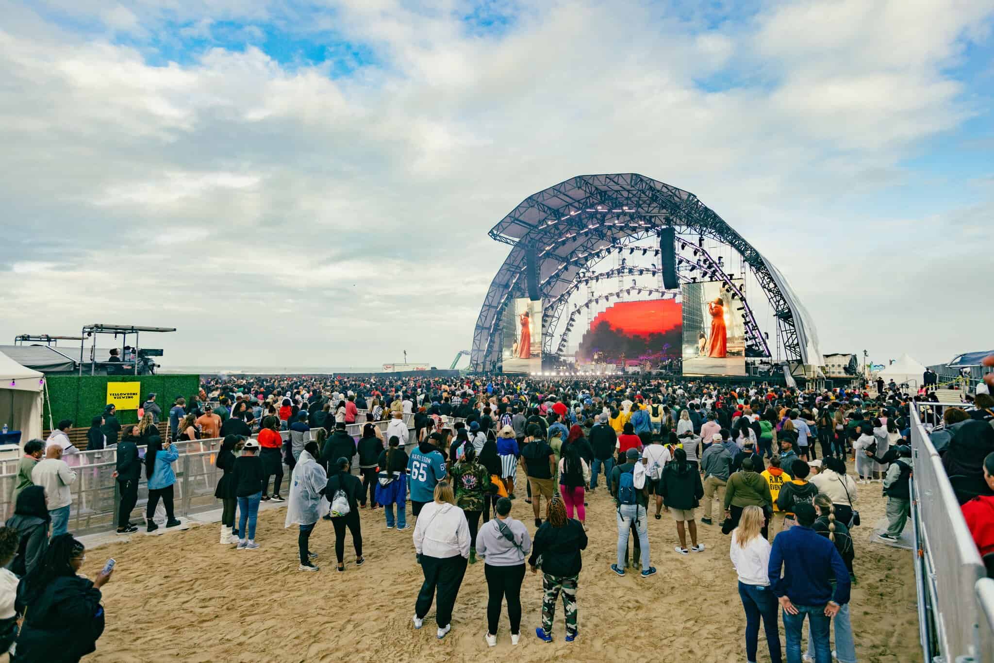Large crowd gathers at the Oceanfront for "Something In The Water" facing a large outdoor concert stage with lights and screens under a partly cloudy sky.