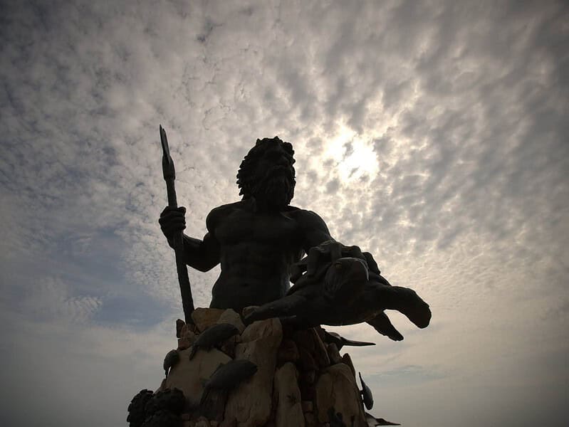 A large statue of a bearded man holding a trident and a sea turtle, known as the King Neptune statue is silhouetted against a cloudy sky.