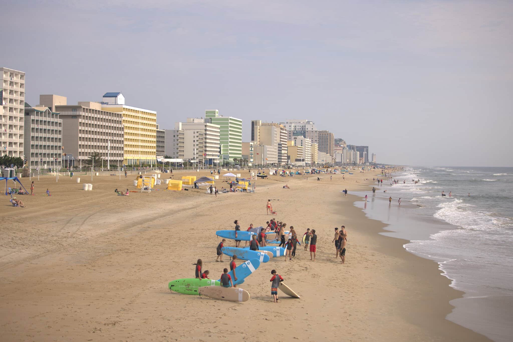 People gather with surfboards on a sandy beach lined with high-rise hotels. Some are swimming, while others walk along the shoreline under a cloudy sky.