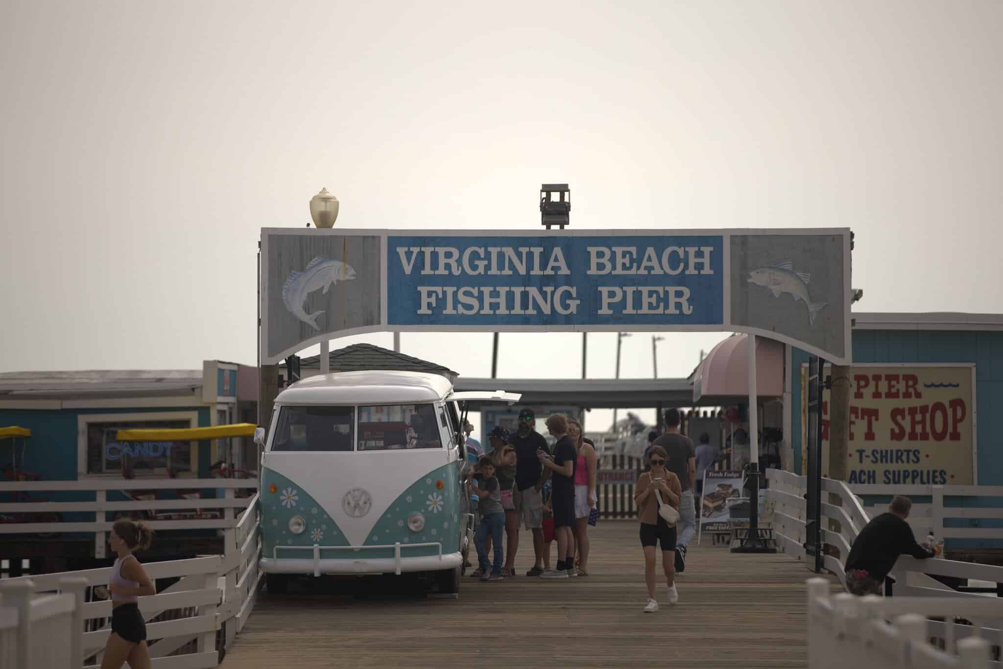People gather at the entrance of the Virginia Beach Fishing Pier, with a vintage Volkswagen van parked nearby and shops visible along the pier.