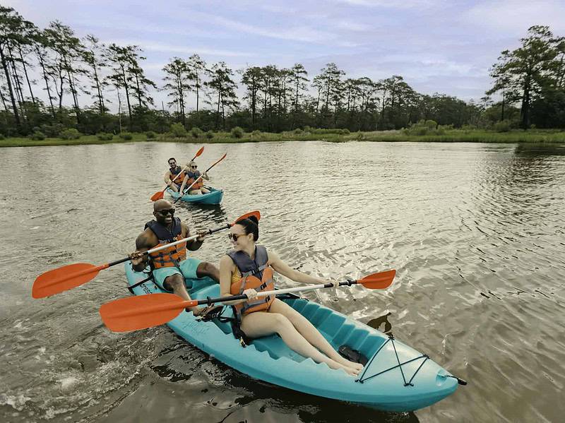 Four people kayaking on a calm river, wearing life vests and using orange paddles. Trees line the riverbank in the background under a cloudy sky.