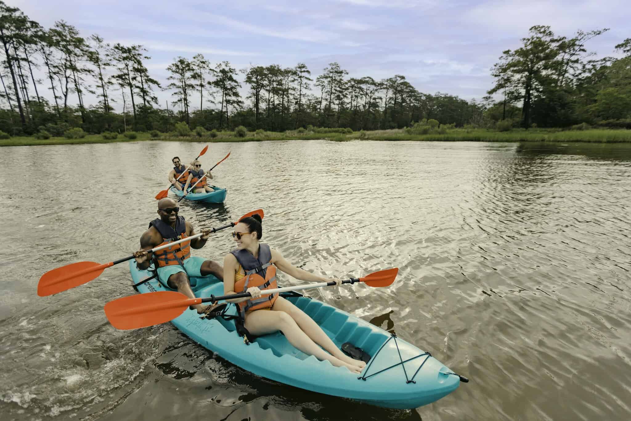 Four people kayaking on a calm river, wearing life vests and using orange paddles. Trees line the riverbank in the background under a cloudy sky.