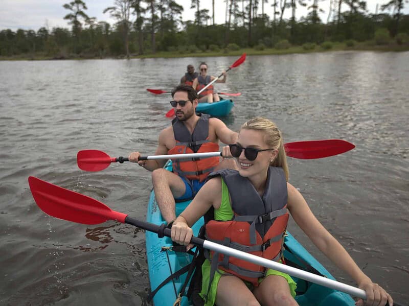Four people wearing life jackets paddle two tandem kayaks at The Narrows, with trees and greenery visible in the background.