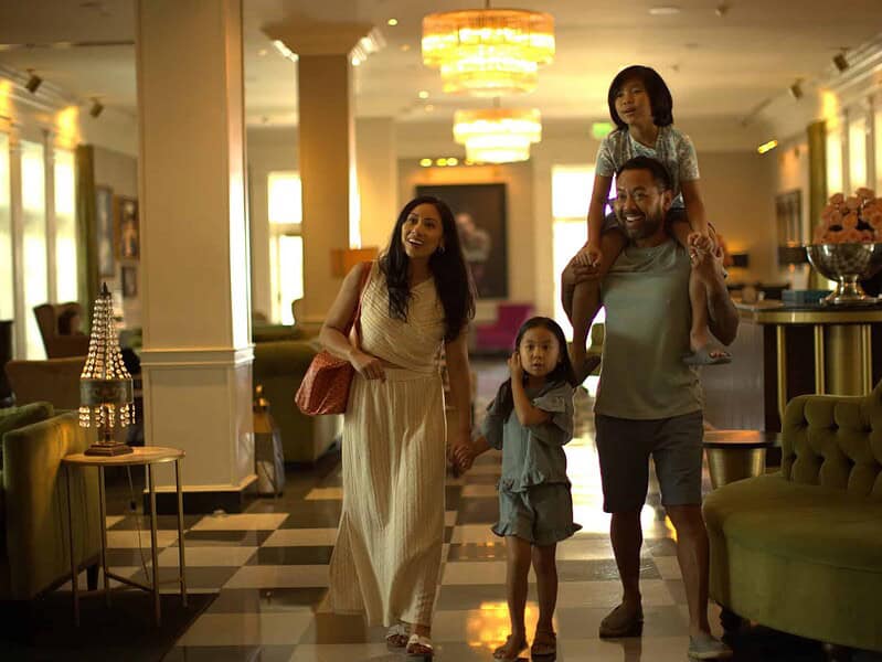 A family of four walks through a hotel lobby; the father carries one child on his shoulders while the mother and the other child walk beside them, all appearing relaxed and happy.