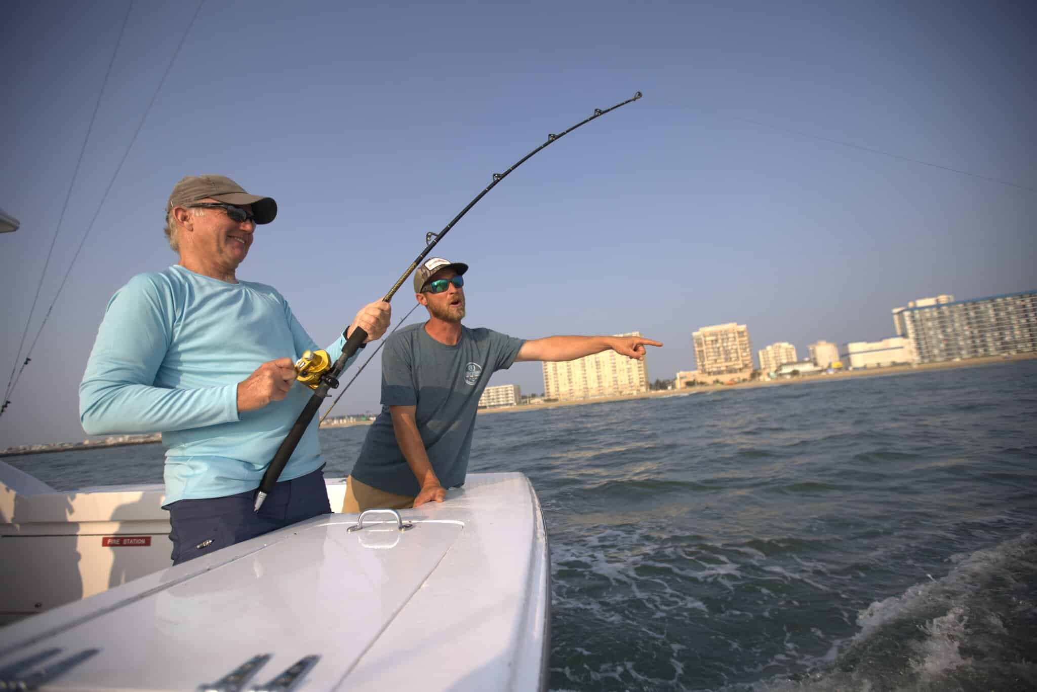 Two men on a boat fishing near Virginia Beach oceanfront; one man holds a fishing rod while the other points toward the water.