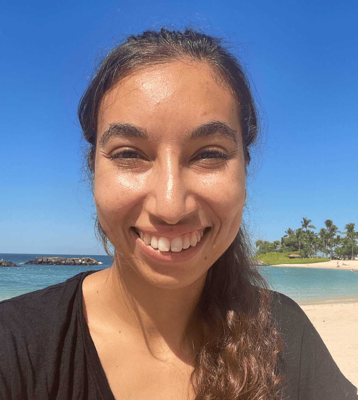 Author Staff member, Sarah Etinas is smiling with long brown curly hair with a beach in the background.