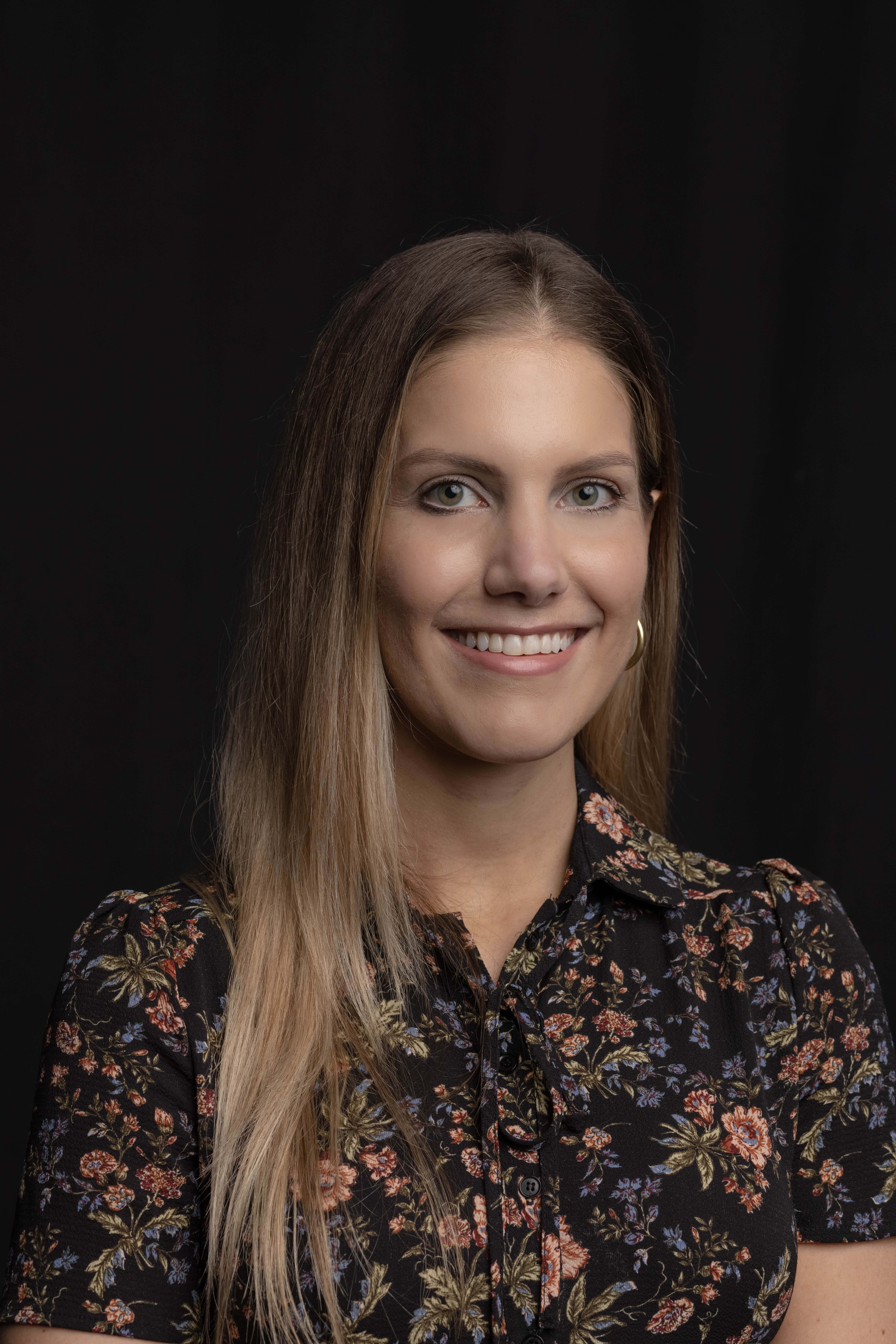 Author Valeria L. Palmertree with long blonde hair smiles, wearing a floral print blouse against a black background.