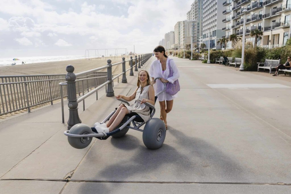 A woman pushes another woman seated in a beach wheelchair along a paved boardwalk by the ocean with tall buildings in the background.