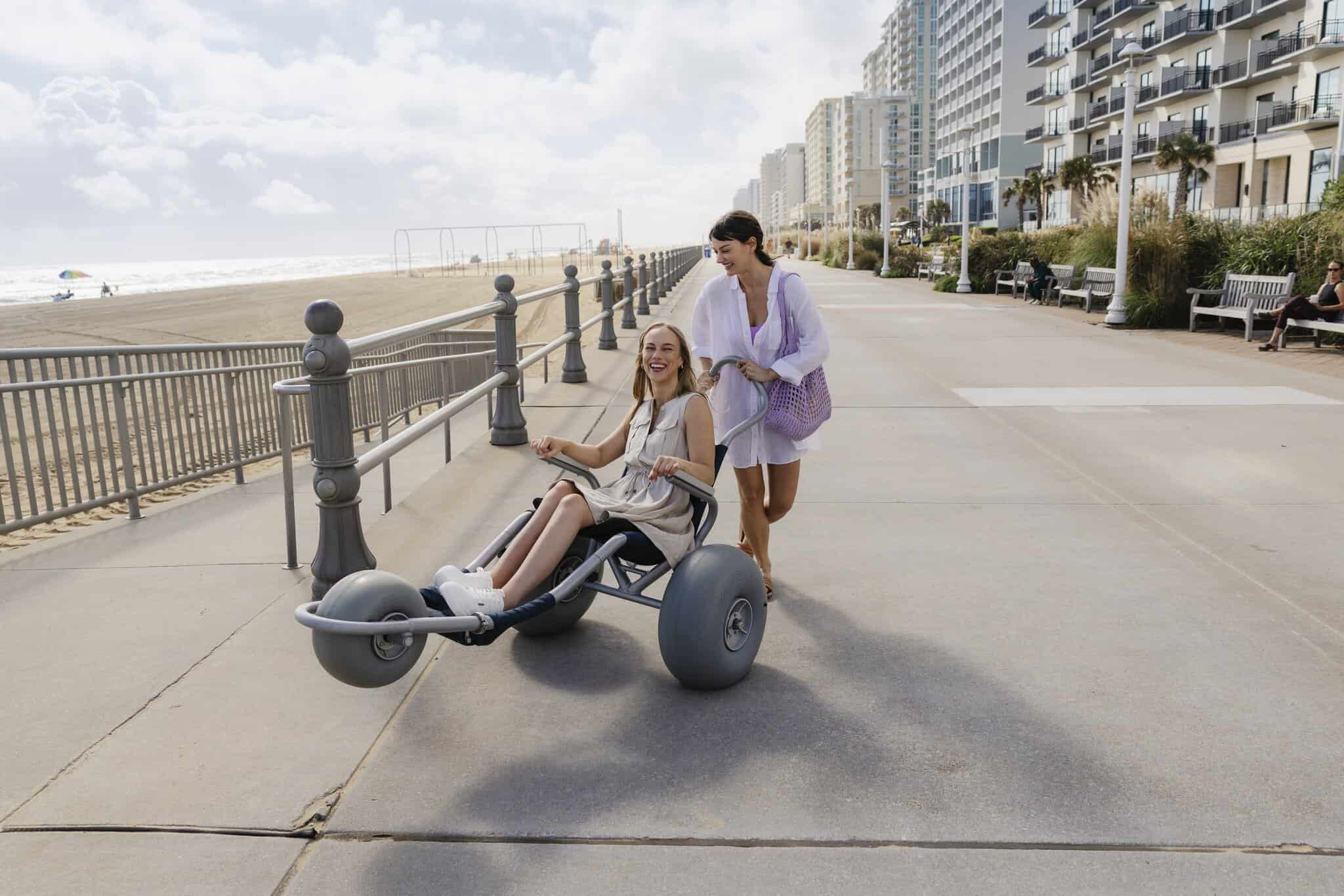 A woman pushes another woman seated in a beach wheelchair along a paved boardwalk by the ocean with tall buildings in the background.