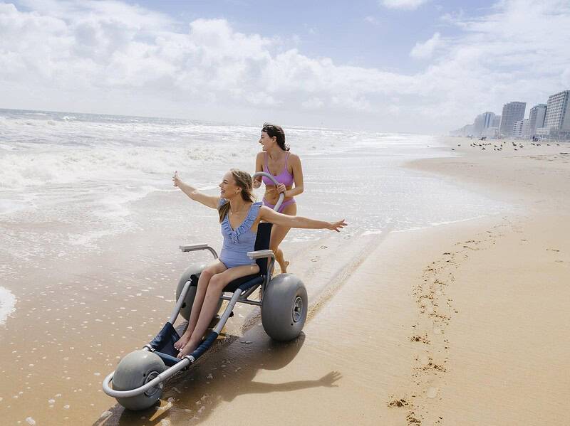 Two women on Virginia Beach; one is sitting in a wheelchair designed for the beach, while the other stands behind her, both facing the ocean with arms outstretched.