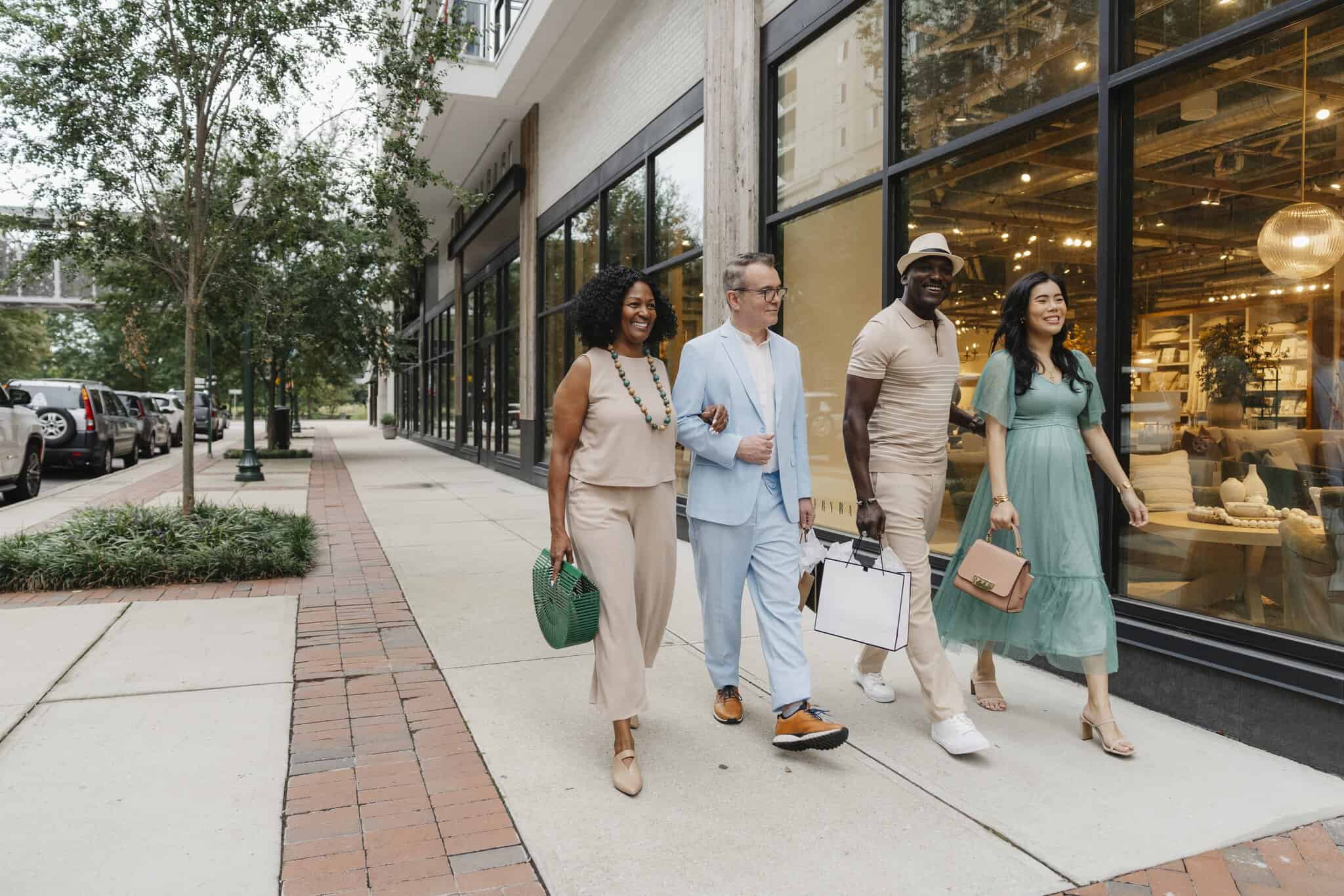 Four adults walk side by side on a city sidewalk, smiling and carrying shopping bags, with storefront windows and trees in the background.