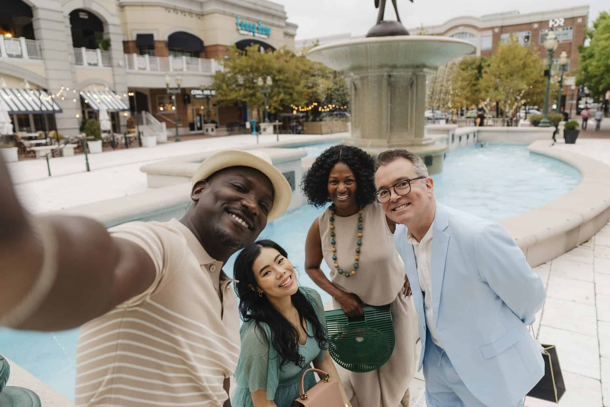 Four people stand together taking a selfie in front of a large fountain in an outdoor shopping center.