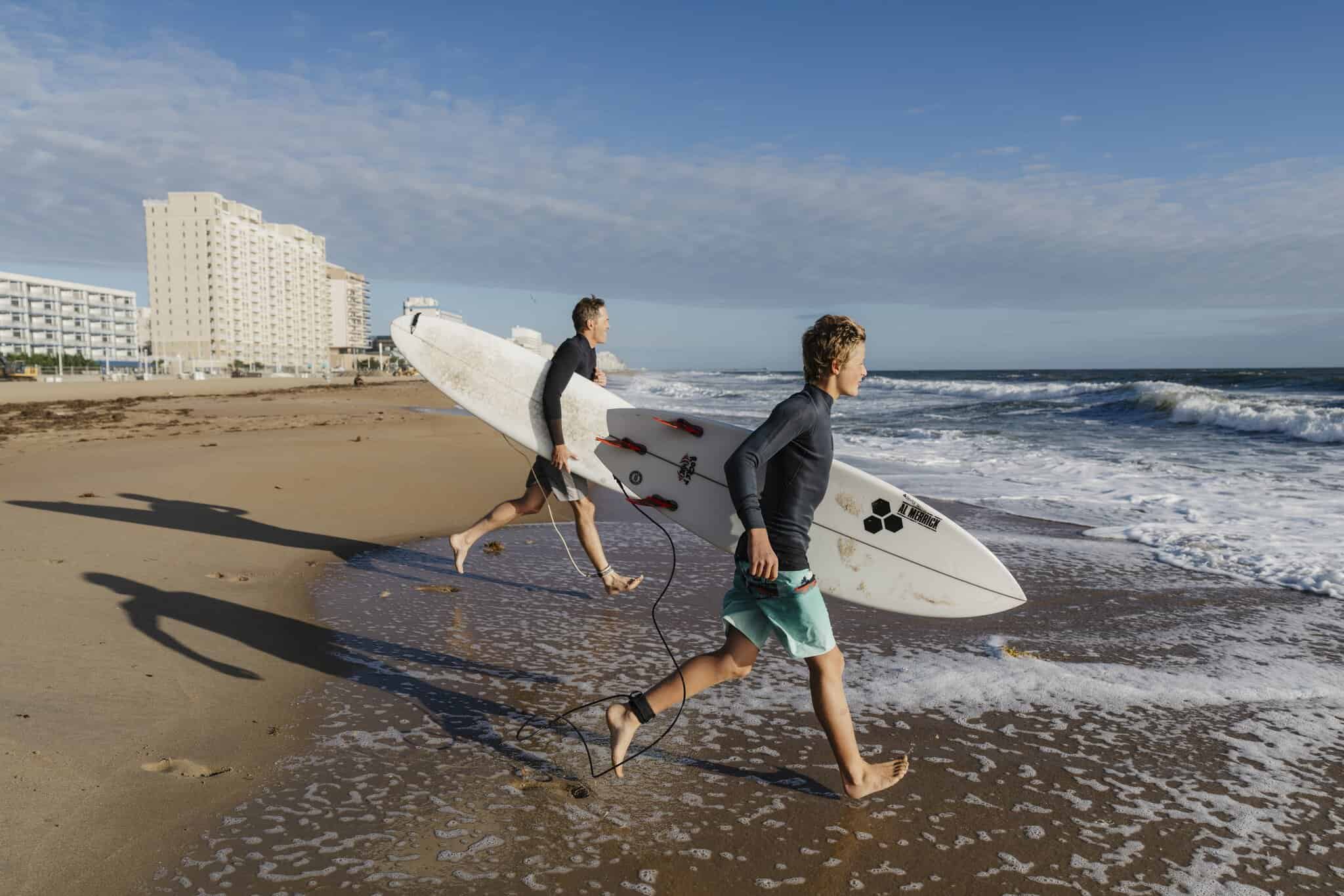 Two people with surfboards run toward the ocean on Virginia Beach, with buildings visible in the background under a partly cloudy sky.