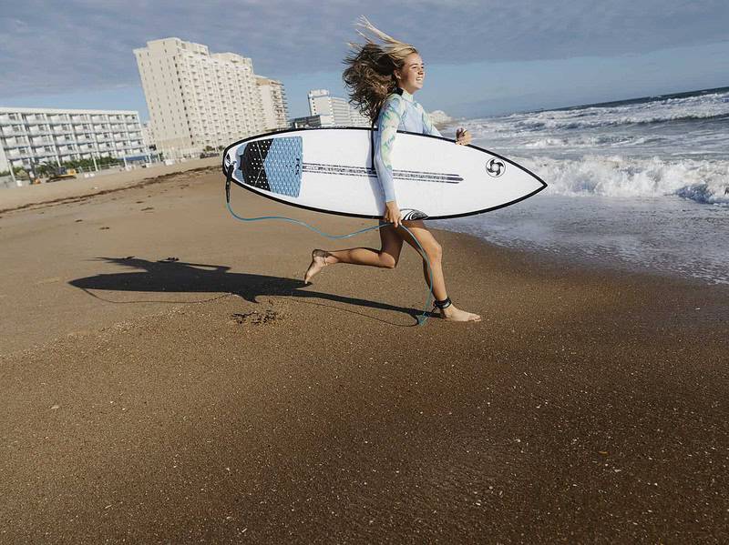 A young girl runs along the oceanfront holding a surfboard, with ocean waves and apartment buildings and hotels visible in the background.