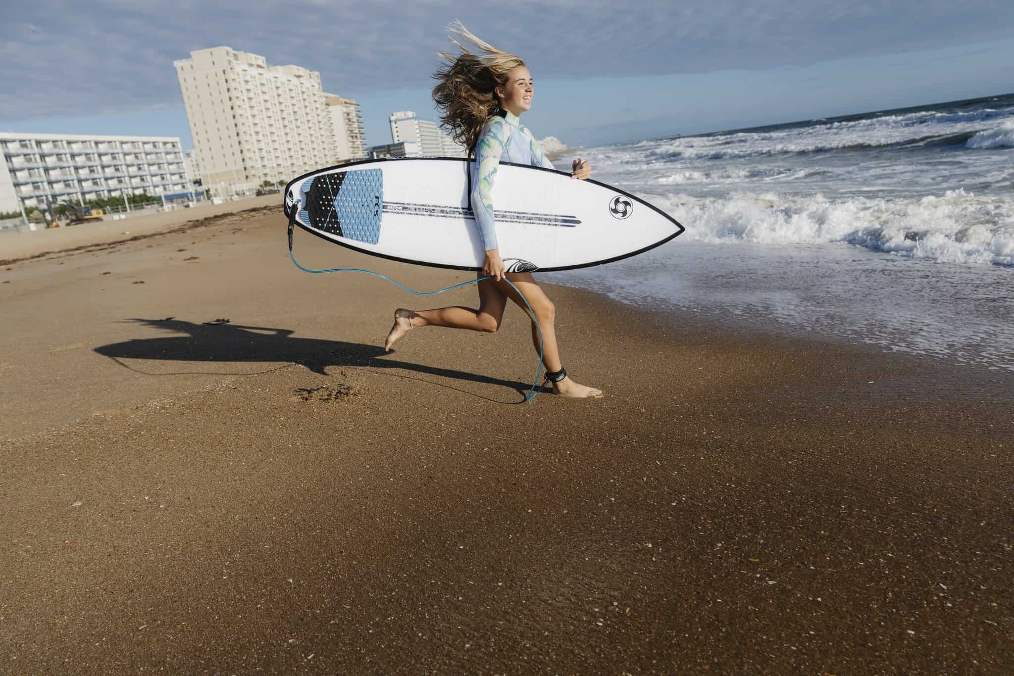 A person runs along a sandy beach holding a surfboard, with ocean waves and tall buildings visible in the background.