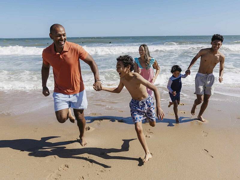 Five people, including three children and two adults, run and smile together on a sandy beach with ocean waves in the background under a clear sky.