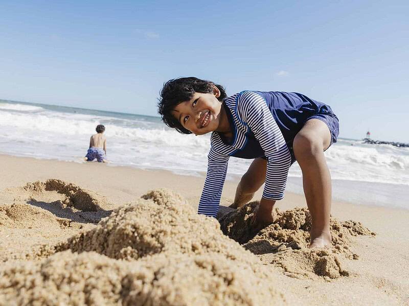 A child in a striped shirt smiles while playing in the sand at the beach, with another child sitting near the shoreline in the background.