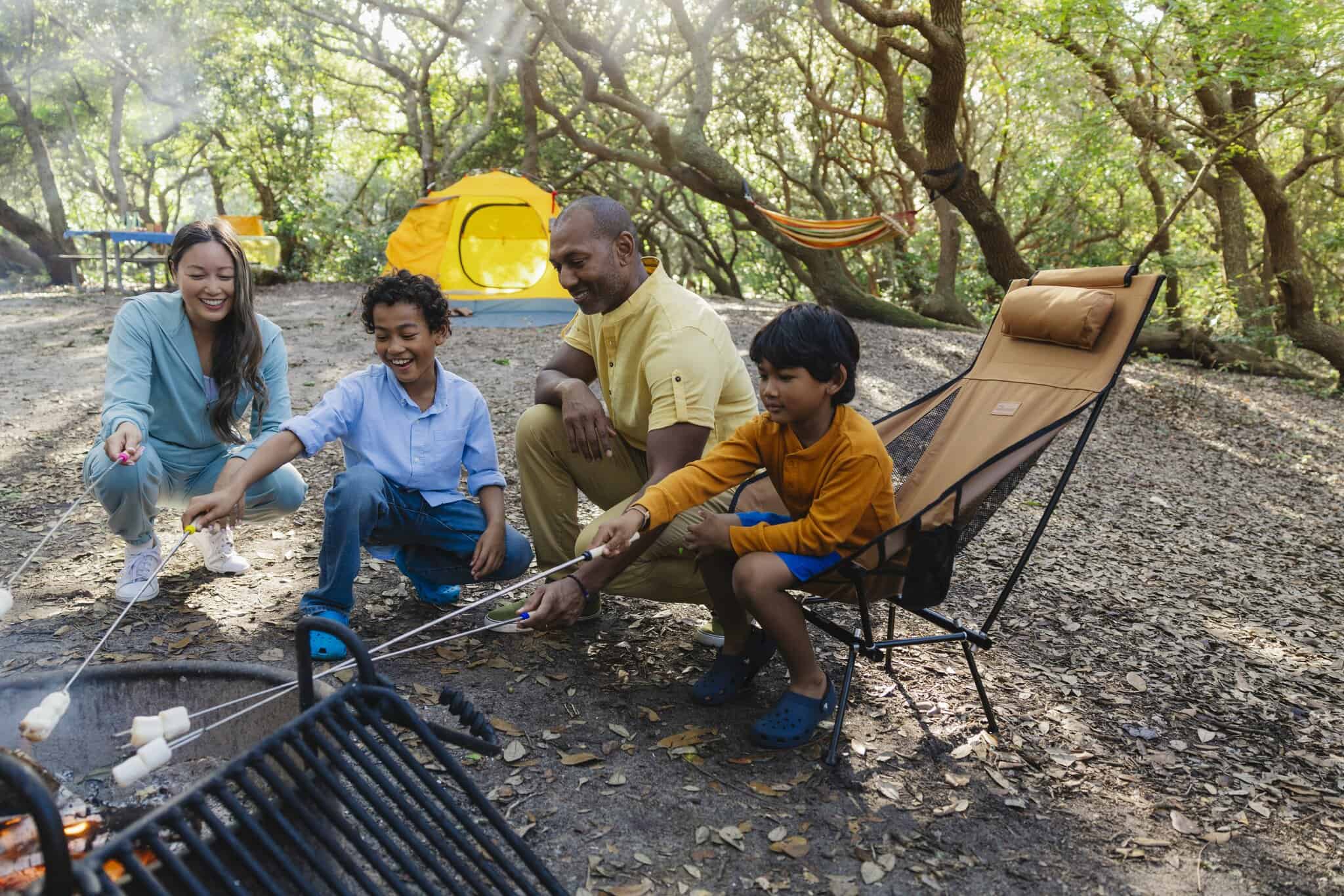 A family of four sits around a campfire at a wooded campsite, roasting marshmallows. A yellow tent and a hammock are visible in the background.