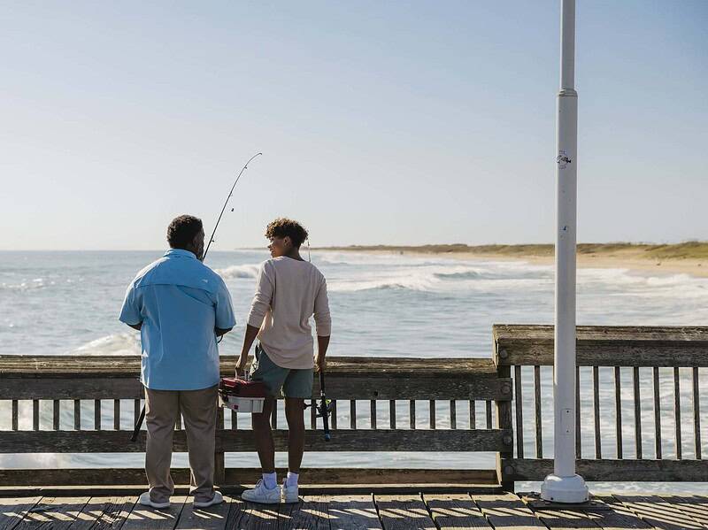 Two people stand on a wooden pier overlooking the ocean; one holds a fishing rod and the other carries a tackle box.