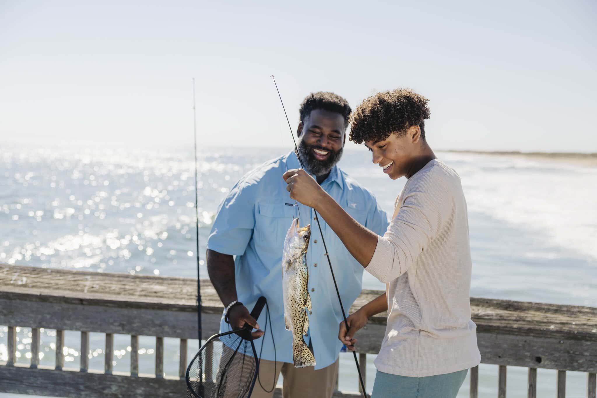 Two people stand on a pier by the ocean, smiling as one holds up a fishing rod with a fish they caught; a fishing net is visible.