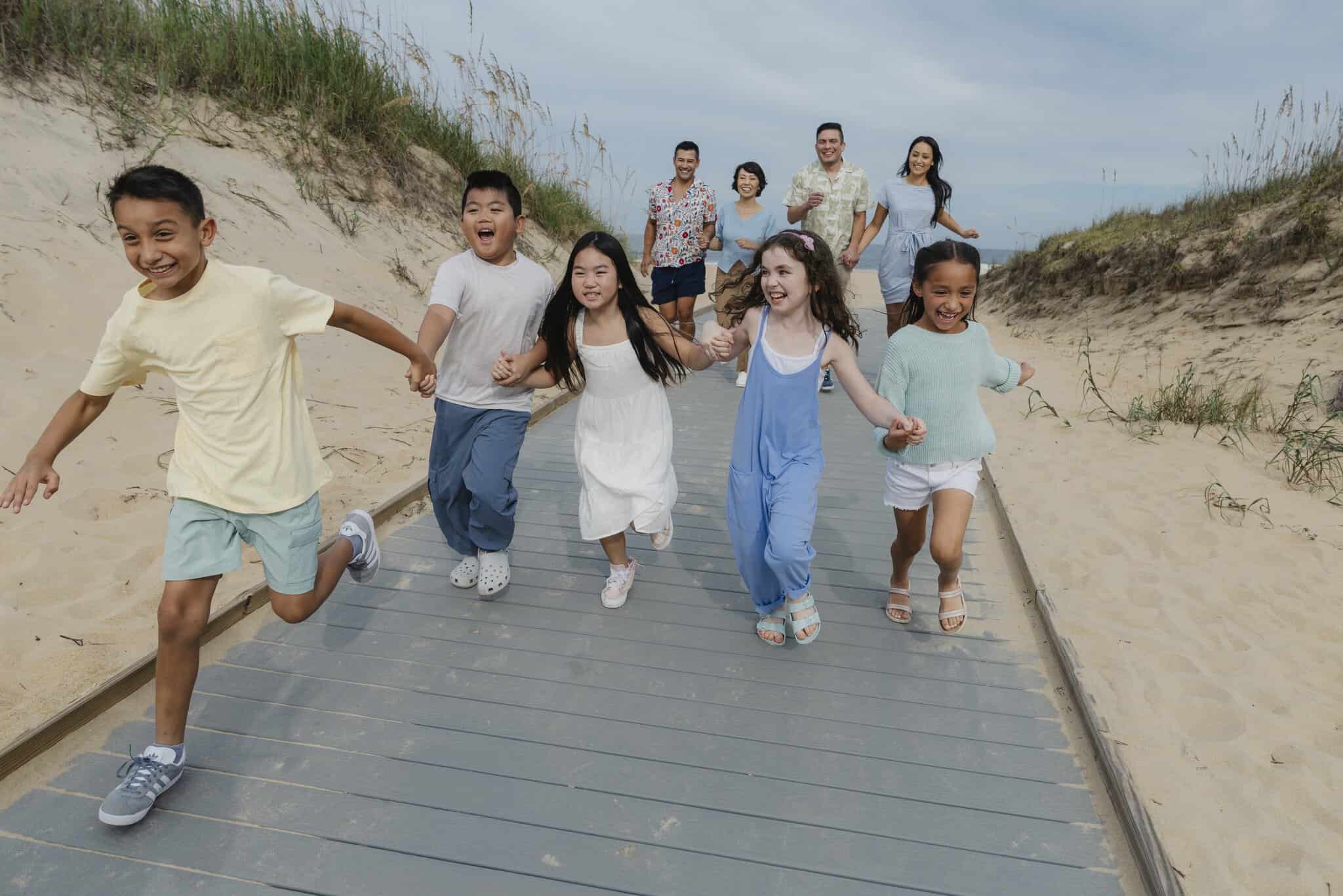 A group of children run on a boardwalk through sand dunes while adults walk behind them, smiling under a partly cloudy sky.