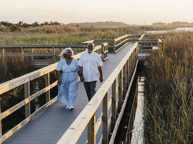An older couple holding hands and walking together on a wooden boardwalk surrounded by tall grass near a body of water at sunset.