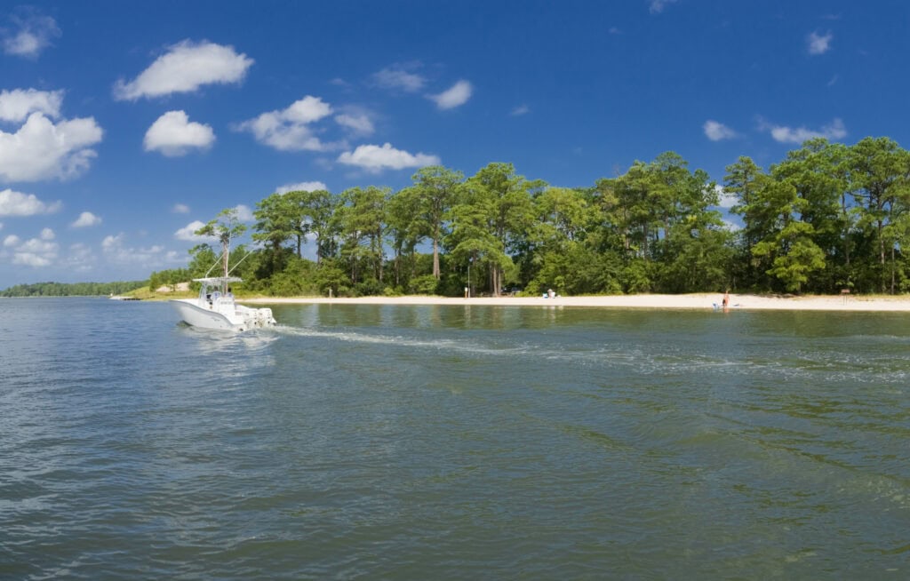 A motorboat navigates through a calm waterway near a sandy beach lined with trees under a bright blue sky.