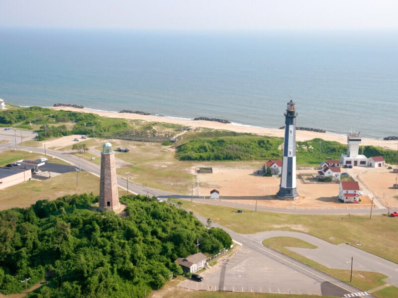Aerial view of two lighthouses near the coast, surrounded by greenery and sandy beach, showcasing a parking area and nearby houses.