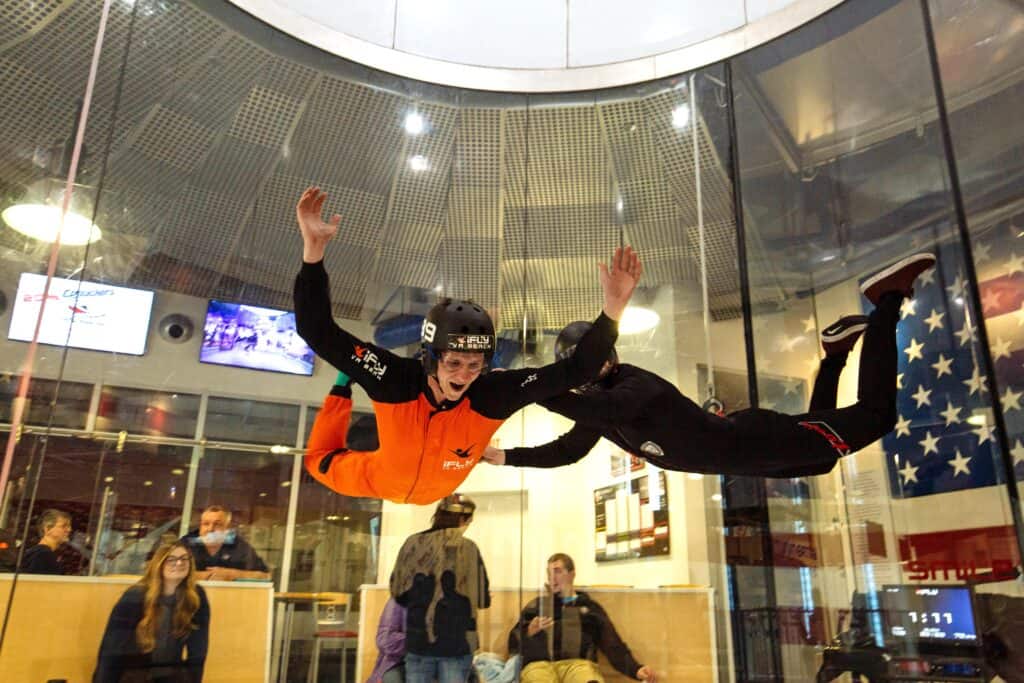 Two skydivers practice indoor skydiving in a wind tunnel, while spectators watch from behind a glass barrier.