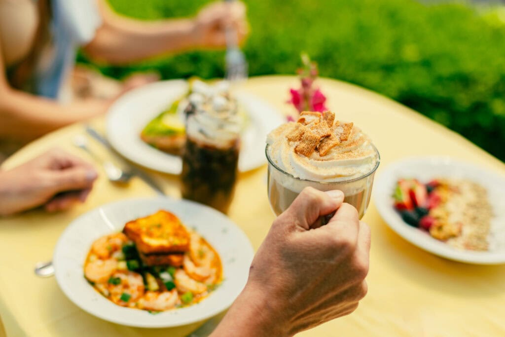 A hand holding a dessert coffee topped with whipped cream and crumbled cookies, with a colorful brunch spread on a yellow table.