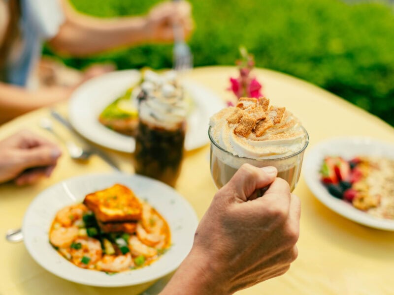 A hand holding a dessert coffee topped with whipped cream and crumbled cookies, with a colorful brunch spread on a yellow table.