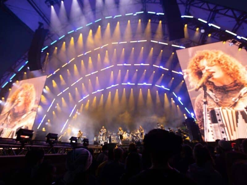 A lively concert scene featuring a band performing on stage with bright lights and large screens displaying a vocalist holding an accordion.