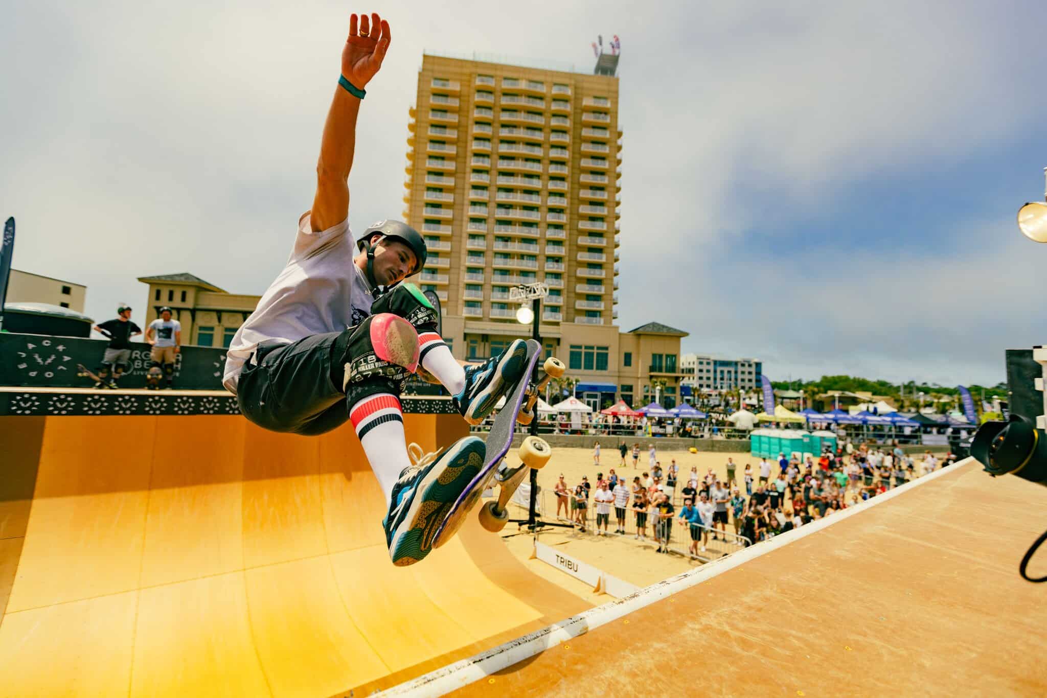 Skateboarder performing an aerial trick on a ramp at a beachside competition, with a crowd and buildings in the background.