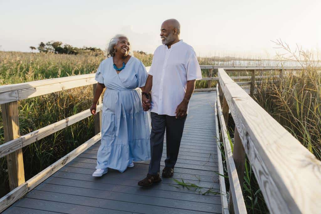 A joyful older couple walks hand in hand along a wooden boardwalk in Back Bay surrounded by tall grass at sunset.