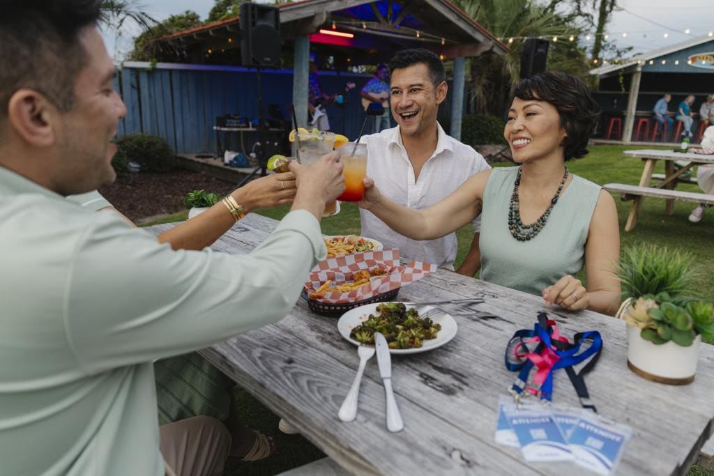 Friends toasting with cocktails at a Baja restaurant with food on a table, greenery and live music in the background.