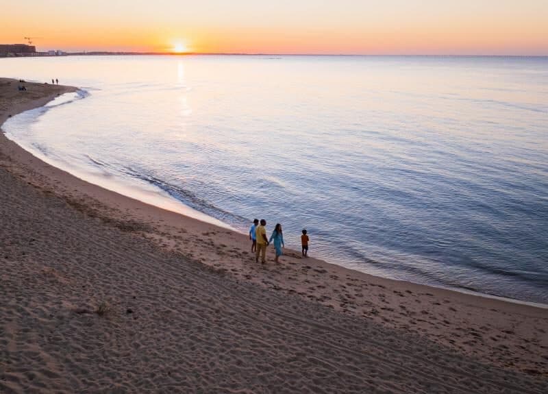 A family walks along a sandy beach at sunset, with calm waves lapping the shore and a vibrant sky in the background.